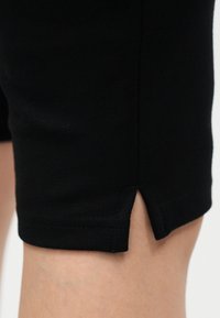 Nærbilde av svarte stoffshorts med liten sideåpning ved kanten over bart kne, mot lys bakgrunn.