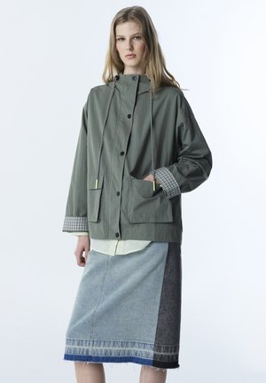 Młoda kobieta ubrana w oversize'ową szaro-zieloną kurtkę z kratkowymi mankietami oraz dwutonową spódnicę jeansową na białym tle.