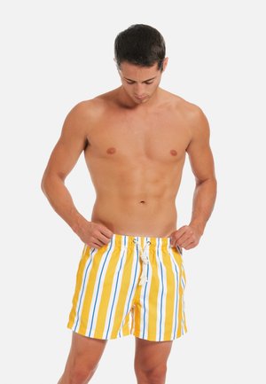 Oberkörperfreier Mann, der gelbe Badeshorts mit weißen und blauen vertikalen Streifen anpasst, steht vor einem schlichten, hellen Hintergrund.