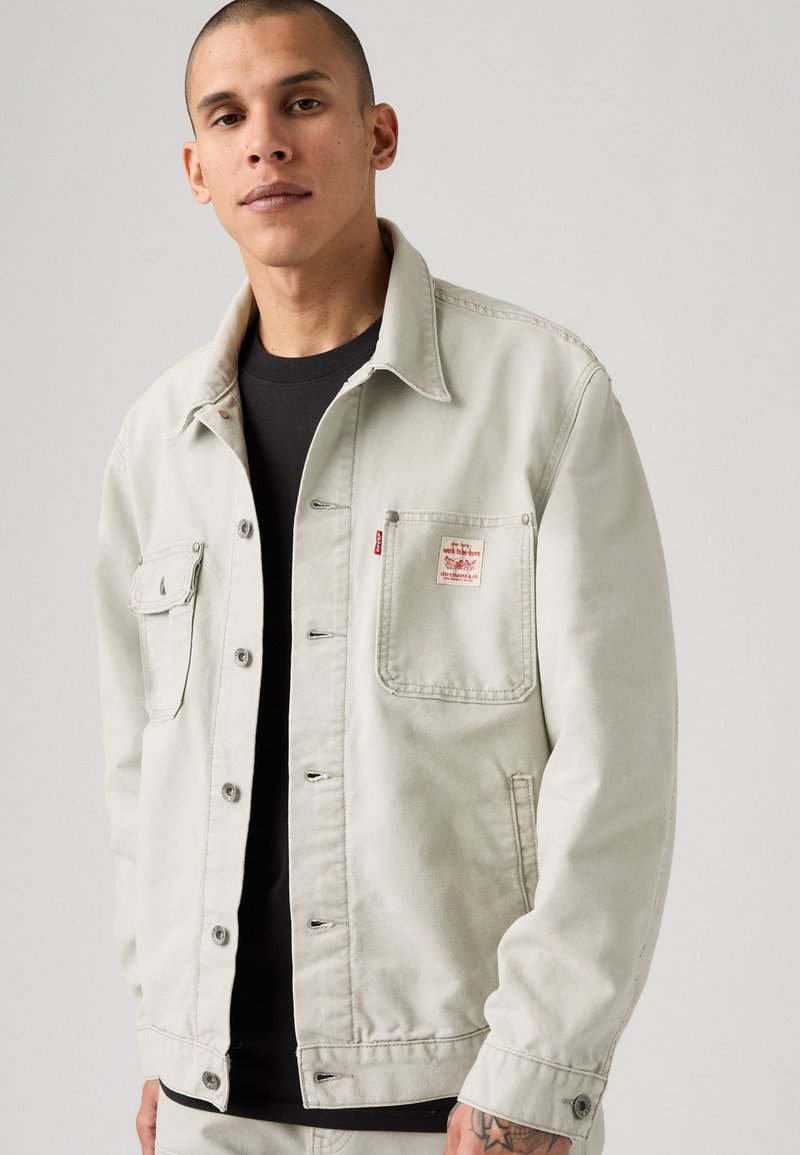 Chaqueta de denim gris claro con cierre de botones, dos bolsillos en el pecho y etiqueta de logo cosida. Presenta un cuello clásico y un corte relajado.