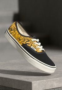 Černé plátěné tenisky se žlutým grafickým designem, bílými šňůrkami a krémovou gumovou podrážkou. Na boku se nachází klasické logo Vans.