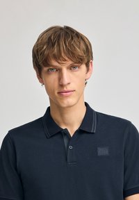 Polo de color marino, confeccionado en tejido texturizado, con cuello abotonado, ribetes en contraste y un parche rectangular con logo en el pecho.