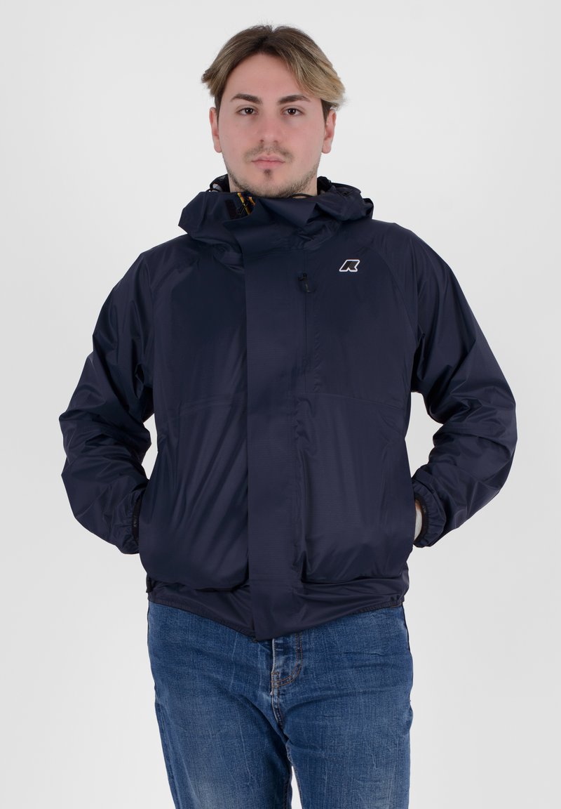 Giacca impermeabile blu navy con cappuccio, chiusura con zip, polsini elasticizzati e un piccolo logo sul petto. Texture liscia, design leggero.