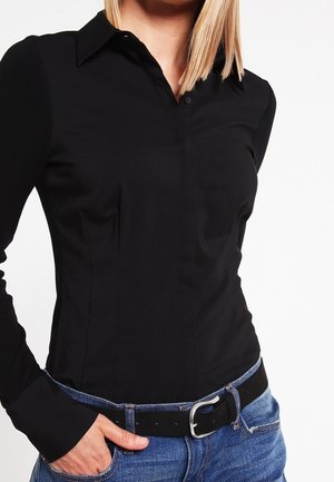 Žena s rovnými blond vlasy má na sobě přiléhavou černou košili na knoflíky, modré džíny a černý pásek, s jednou rukou v kapse.