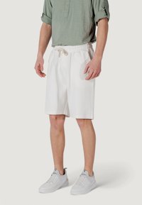 Un uomo è in piedi indossando una camicia verde chiaro e pantaloni corti bianchi, abbinati a scarpe da ginnastica bianche, con le mani rilassate ai lati. Sfondo neutro.