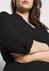 Femme aux bras croisés portant un haut noir à manches longues avec des détails de boutons et des boucles d’oreilles en argent en forme de créoles sur un fond uni.