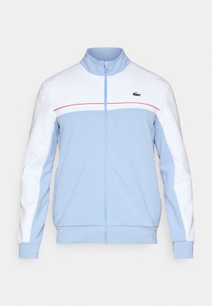 Lichtblauwe ritsjack met witte mouwen, rode accentlijn en een groen logo op de borst. Gemaakt van een zacht, ademend materiaal.