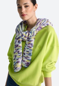 A színes fonalból kötött sál lila, sárga és kék mintákkal, egy élénk zöld, nagy méretű pulóverre van átvetve.
