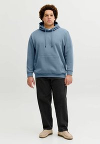 Modra jopa z kapuco v sproščenem kroju, s kenguru žepom, kapuco z vrvico in rebrastimi manšetami, kombinirana s temnimi hlačami in bež čevlji.