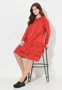Rotes Kleid mit einem einfarbigen Oberteil und einem strukturierten, bestickten Spitzenrock mit gewellten Kanten, kombiniert mit schwarzen Slippern.
