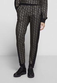Pantalones de chándal negros con un patrón de logotipos EA7 dorados repetidos, corte ajustado, cinturilla elástica y bolsillos laterales. Combinados con zapatillas negras.