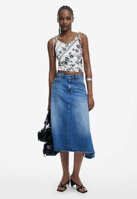 Weißes Crop-Top mit schwarzem Blumenmuster, kombiniert mit einem blauen Denim-Midirock. Das Model hält eine schwarze Tasche und trägt schwarze sandalen mit Riemchen.