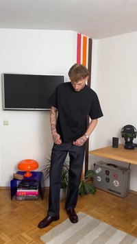 Camiseta negra de manga corta, pantalones de mezclilla oscuros y zapatos burdeos. Tatuajes notables en los brazos y accesorios que incluyen un reloj y gafas.