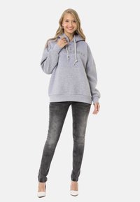 Hellgrauer übergroßer Kapuzenpullover mit Kordeln und gesticktem Logo, kombiniert mit dunklen Jeans und weißen Absätzen, stehend in einem neutralen Raum.