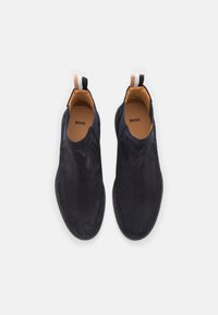 Chaussures slip-on en nubuck bleu marine avec une texture lisse, semelle en caoutchouc noir et accents intérieurs beige. Présente un tirant noir et blanc.