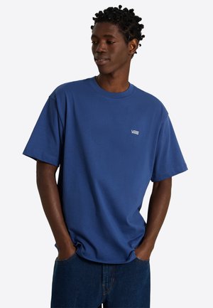 Mann mit Händen in den Taschen, der ein blaues Vans-T-Shirt und dunkelblaue Jeans trägt und leicht zur Seite mit neutralem Gesichtsausdruck schaut.