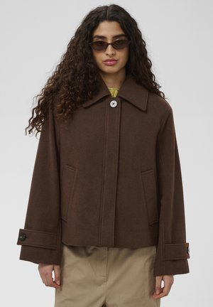 Vrouw met lang krullend haar, donkere zonnebril, een bruine jas met knopen en beige broek, staand tegen een effen achtergrond.