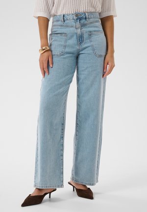 Femme portant un jean bleu clair à jambes larges avec poches plaquées à l'avant, un haut en tricot beige, des talons marron pointus et un gros bracelet en or.