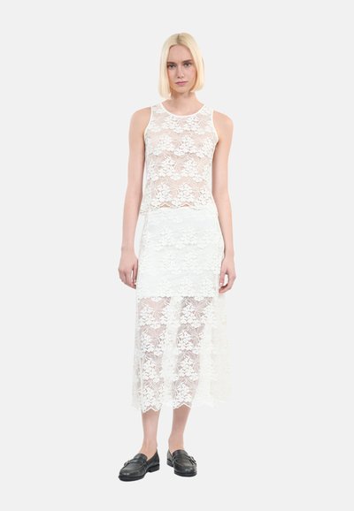 Witte kanten twee-delige set met een mouwloze top en een vloerlange rok met gelaagde kanten patronen, gecombineerd met zwarte loafers.