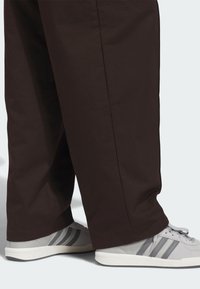 Καφέ παντελόνια με φαρδιά μπατζάκια συνδυασμένα με γκρι αθλητικά παπούτσια που έχουν μαύρες ρίγες και  ανοιχτούς τόνους. Το υλικό φαίνεται λείο με ελάχιστη υφή.