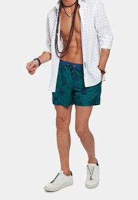 Bañador corto en verde azulado con patrones de hojas oscuras, combinado con una camisa blanca de lunares y zapatillas blancas con detalles de cremallera.