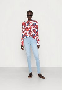 Camisa com botões de padrão floral em vermelho e azul, combinada com jeans skinny azul claro e chinelos pretos, apresentada sobre um fundo neutro.