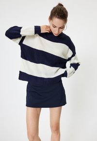 Gebreide trui met marineblauwe en crèmekleurige horizontale strepen, met een hoge halslijn en geribbelde manchetten. Gepaard met een marineblauwe minirok.