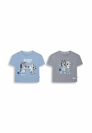 Smiling young boy with blonde hair wearing a grey cartoon character t-shirt, blue jeans, and grey sneakers, standing against a plain background.