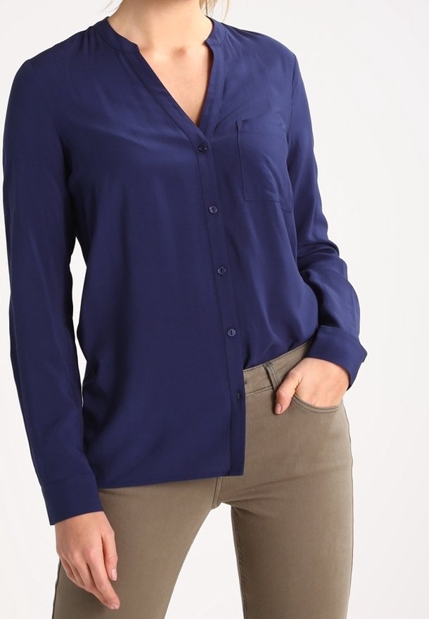 Chemise bleu marine à boutons en tissu lisse avec col montant, poche poitrine unique et manches longues, associée à un pantalon beige.