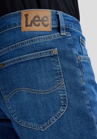 Niebieskie dżinsy z denimu, zbliżenie na tylną kieszeń z charakterystycznym przeszyciem oraz brązową skórzaną naszywką marki Lee na pasku.