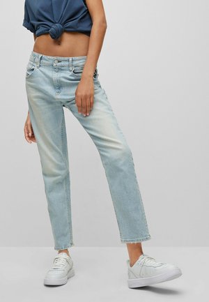 Persoon draagt lichtblauwe rechte jeans, witte sneakers en een donkerblauw geknoopt cropped shirt tegen een effen achtergrond.