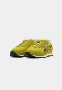 Žute Reebok tenisice s crnim logotipom i trakama, bijelim srednjim potplatima, crnim potplatima i podstavom s amortizacijom, prikazane jedna pored druge na bijeloj pozadini.