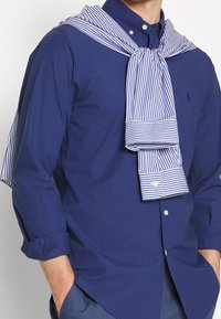 Camicia blu navy con bottoni e maniche lunghe, con una cravatta a righe bianche e blu appesa sulle spalle.