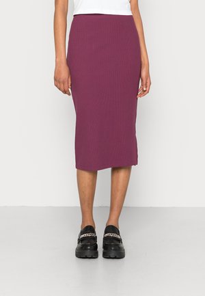 Kvinde iført en tætsiddende, knælange lilla nederdel med sorte loafers med sølvkæde detaljer, stående mod en ensfarvet baggrund.