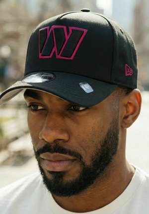 Mann mit Bart, der eine schwarze Mütze mit einem großen, lila umrandeten "W"-Logo und ein weißes Hemd trägt und im Freien leicht nach unten schaut.