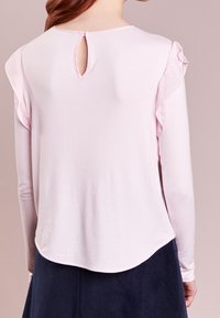 Blouse à manches longues rose clair avec des accents de volants aux épaules et une ouverture en forme de goutte d'eau au dos. Texture douce avec un ourlet arrondi.