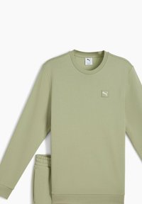 Lys grøn sweatshirt med rund halsudskæring og lange ærmer, der har et lille broderet logo på brystet og en elastisk talje på bukserne.