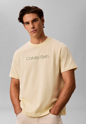 Młody mężczyzna z brązowymi włosami, ubrany w jasnżółtą koszulkę Calvin Klein i jasne spodnie, pozujący z rękami w kieszeniach na szarym tle.