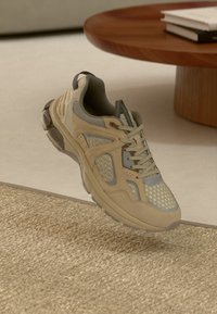 Sapatilha desportiva bege e cinza com detalhes em malha e camurça, atacadores atados, posicionada sobre um tapete texturizado perto de uma mesa redonda de madeira.