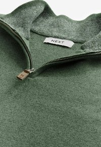 Grüner Zip-Pullover mit einer strukturierten, weichen Oberfläche, der einen hohen Kragen und einen Metallreißverschluss mit einem Lederzieher hat. Markenetikett sichtbar.