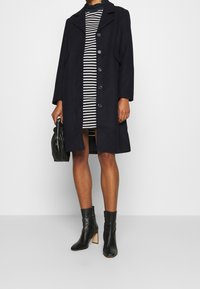 Navy väiri villasegu mantel, millel on nurgaga krae ja nööpidega esikülg, kombineeritud triibulise kleidi ja mustade nahast pahkläbimõõtmetega saapaga. Aksessuaarideks on must kott.