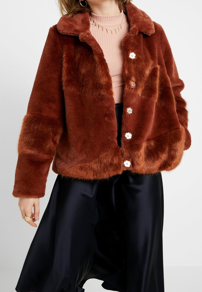 Femme portant une veste en fausse fourrure couleur rouille sur un haut côtelé rose pâle et un pantalon large en satin noir, accessoirisée avec des bijoux en or.