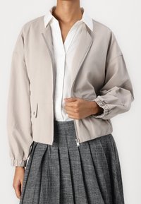 Chaqueta bomber corta de color gris claro con cremallera frontal, puños elásticos y bolsillos laterales, combinada con una camisa blanca y una falda gris con pliegues.