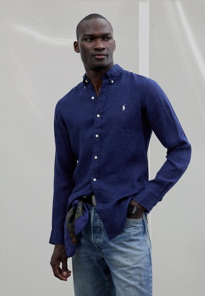 Marineblauw linnen overhemd met lange mouwen en een button-down kraag, met een wit geborduurd logo en een blauw patroonaccessoire gestrikt in de taille.