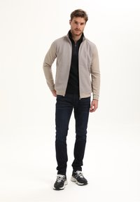 Beżowa kurtka zapinana na zamek z textured przodem, czarnym wysokim kołnierzem, warstwowym krojem. Nosi się ją na ciemnej koszuli, w połączeniu z ciemnymi dżinsami i czarnymi trampkami.