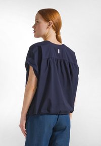 Frau mit roten Haaren in einem Pferdeschwanz, die eine lockere marineblaue kurzärmelige Bluse und blaue Jeansshorts trägt, steht leicht seitlich gewandt.