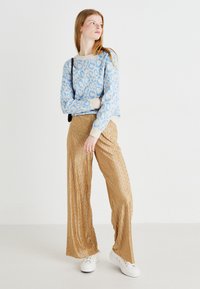 Ανοιχτό μπλε πουλόβερ με λεοπάρ σχέδιο και κρεμ μανίκια, συνδυασμένο με χρυσά πλισέ παντελόνια πλατιάς γραμμής και λευκά αθλητικά παπούτσια.