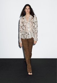 Blouse en crêpe crème avec des taches brunes, boutonnée à l'avant ; associée à un pantalon évasé en daim marron et des sandales noires.