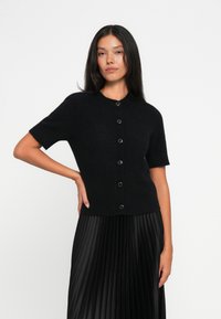 Cardigan corto nero a maniche corte realizzato in materiale morbido, con scollatura rotonda e sei bottoni neri sul davanti.