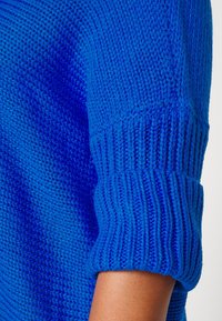Stickad blå tröja med ribbad ärmslut, som visar ett strukturerat, öppet stickat mönster och en åtsittande passform runt armen.